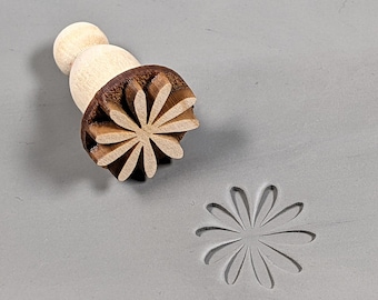May include: A wooden stamp with a brown daisy design. The stamp is used to create an impression in clay or dough.