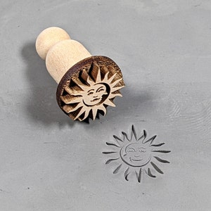 May include: A wooden stamp with a handle and a carved design of a sun with a face. The stamp is used to create an impression of the sun on clay or other materials.