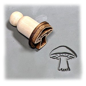 May include: A wooden mushroom stamp with a brown handle and a detailed mushroom design. The stamp is used to create an impression of a mushroom on a surface.