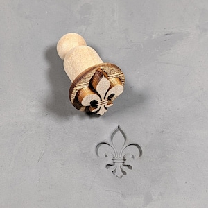 May include: A wooden stamp with a fleur-de-lis design. The stamp is used to create an impression of the design on a surface.
