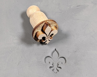 May include: A wooden stamp with a fleur-de-lis design. The stamp is used to create an impression of the design on a surface.