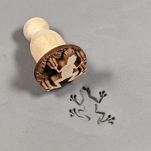 May include: A wooden stamp with a frog silhouette design. The stamp is used to create an impression of a frog on clay or dough.