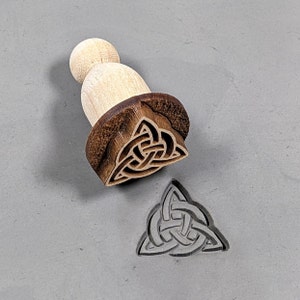 May include: A wooden stamp with a Celtic knot design. The stamp is in the shape of a triangle and has a brown wooden handle. The stamp is used to create impressions in clay or dough.