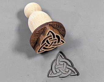 May include: A wooden stamp with a Celtic knot design. The stamp is in the shape of a triangle and has a brown wooden handle. The stamp is used to create impressions in clay or dough.