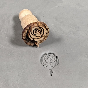 May include: A wooden rose-shaped stamp with a brown handle. The stamp is used to create an impression of a rose in clay or dough.