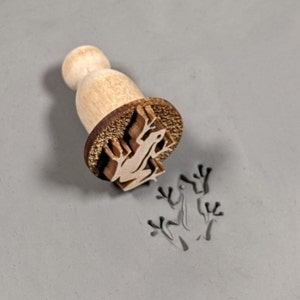 May include: A wooden stamp with a frog design. The stamp is being used to create an impression of a frog in clay.