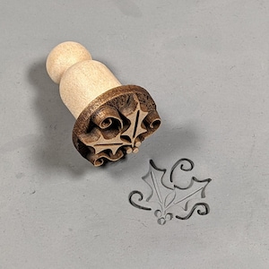 May include: A wooden stamp with a holly leaf design. The stamp is brown and has a wooden handle. The stamp is used to create an impression of a holly leaf on clay or dough.