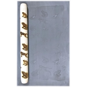 May include: A wooden rolling pin with a pattern of frogs burned into the surface. The rolling pin is next to a gray surface with a repeating pattern of embossed frogs.