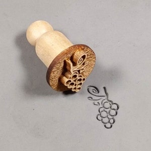 May include: A wooden stamp with a grape design. The stamp is used to create an impression of a grape cluster on clay or dough.