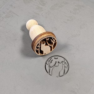 May include: Wooden stamp with a brown handle and a circular design of a cat in a forest. The design is carved into the wood and is visible on the stamp.