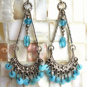 May include: Silver dangle earrings with a crescent moon design and blue glass beads. The earrings have a layered look with multiple strands of beads.