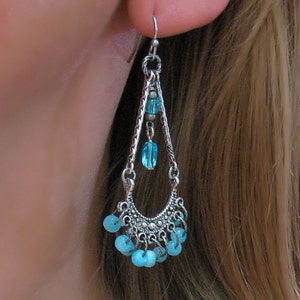 May include: Silver dangle earrings with a teardrop shape and blue beads. The earrings have a silver chain with a small blue bead at the top and a larger blue bead at the bottom. The earrings are accented with a silver crescent moon shape at the bottom.