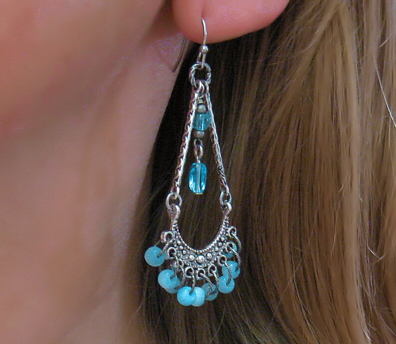 May include: Silver dangle earrings with a teardrop shape and blue beads. The earrings have a silver chain with a small blue bead at the top and a larger blue bead at the bottom. The earrings are accented with a silver crescent moon shape at the bottom.