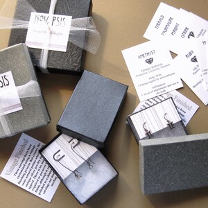 May include: A collection of small, gray gift boxes with white ribbon and labels. The boxes contain earrings and are labeled with the text "NOV, PSIS". There are also several small white cards with text about different gemstones.