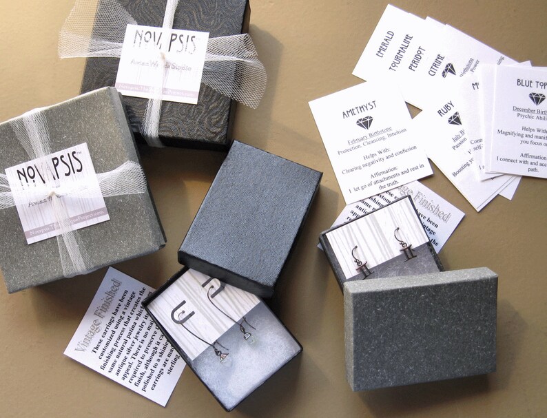 May include: A collection of small, gray gift boxes with white ribbon and labels. The boxes contain earrings and are labeled with the text "NOV, PSIS". There are also several small white cards with text about different gemstones.