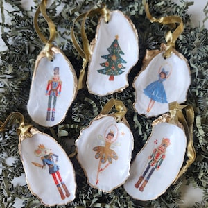 May include: Six oyster shell Christmas ornaments with gold trim and ribbon hangers. Each shell features a different festive design, including a Christmas tree, a ballerina, and a nutcracker soldier.