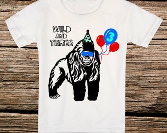 May include: A white short-sleeve t-shirt featuring a black and white graphic of a gorilla wearing a teal polka-dot party hat and blue sunglasses. The text "WILD AND THREE" is printed above the gorilla, accompanied by two red balloons and one blue balloon with the number "3" inside.