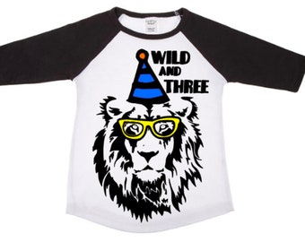 May include: A white raglan t-shirt with black three-quarter length sleeves featuring a graphic of a lion wearing yellow glasses and a blue and black striped party hat with an orange pom-pom, accompanied by the black text WILD AND THREE.