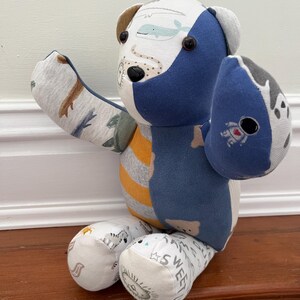 May include: A patchwork teddy bear made from various patterned fabrics. The bear has blue, white, grey, and yellow sections. One arm features an astronaut print, the other a dinosaur print. The bear's feet have the word "SWEET" printed on them.