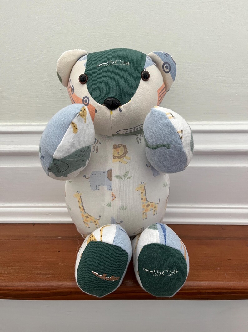 May include: A handmade patchwork bear toy. The bear has a green and cream-coloured head with black button eyes and a black nose. The body is white fabric printed with giraffes, elephants, and lions. The arms and legs are blue and green.
