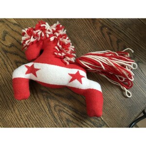 Puede incluir: Juguete de peluche de caballo rojo y blanco con una franja blanca y estrellas rojas. El caballo tiene una cola larga hecha de hilo rojo y blanco.
