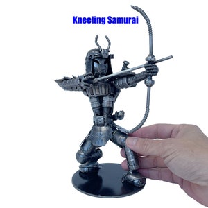 Puede incluir: Una escultura de metal de un guerrero samurái arrodillado sosteniendo un arco y una flecha. La escultura está hecha de metal reciclado y tiene un aspecto rústico e industrial.