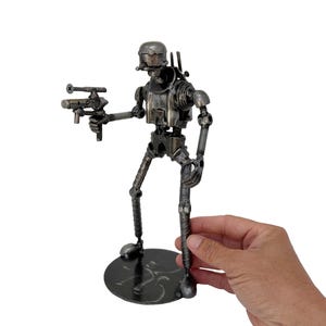 May include: A metallic robot figurine constructed from recycled hardware components, including screws, bolts, and metal rods. The robot features a rounded head, a torso with visible mechanical parts, and articulated limbs, holding a small blaster.