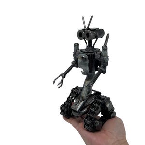 May include: A metallic robot sculpture constructed from recycled hardware components including nuts, bolts, screws, and metal chains. The figure features two circular eyes, a pair of thin arms with claw-like hands, and a base made of tank-style treads.