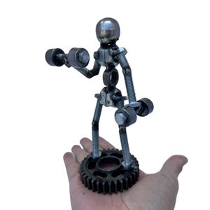 Könnte beinhalten: Eine Metallskulptur einer Person, die Gewichte hebt. Die Skulptur besteht aus Muttern, Schrauben und anderen Metallteilen. Die Person steht auf einem Zahnrad.