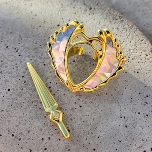 May include: A gold-tone heart-shaped ring with iridescent pink and blue accents. The ring is accompanied by a gold-tone sword-shaped charm with a clear rhinestone.