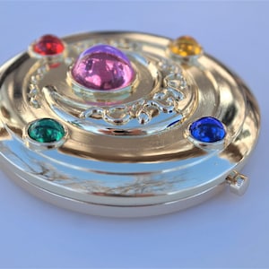 May include: A gold-colored compact mirror with a crescent moon design and five colorful gemstones. The gemstones are red, pink, green, yellow, and blue.