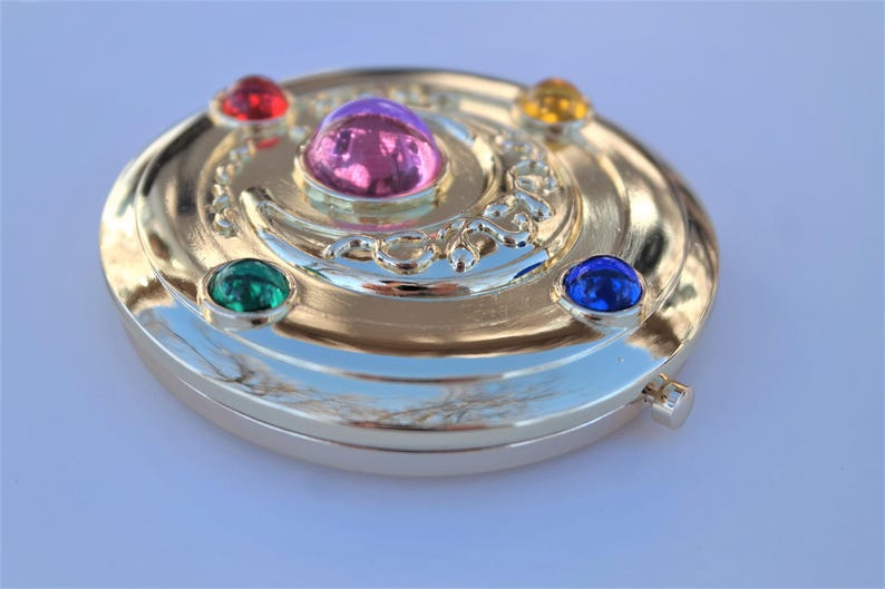 May include: A gold-colored compact mirror with a crescent moon design and five colorful gemstones. The gemstones are red, pink, green, yellow, and blue.