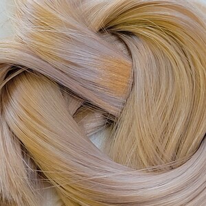 May include: A close-up of a bundle of light blonde hair with a slight golden hue. The hair is soft and silky, with a subtle sheen.