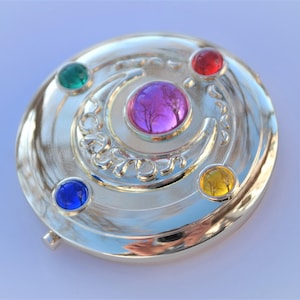May include: A silver-colored compact mirror with a crescent moon design and five colorful gems. The gems are red, green, blue, yellow, and pink.