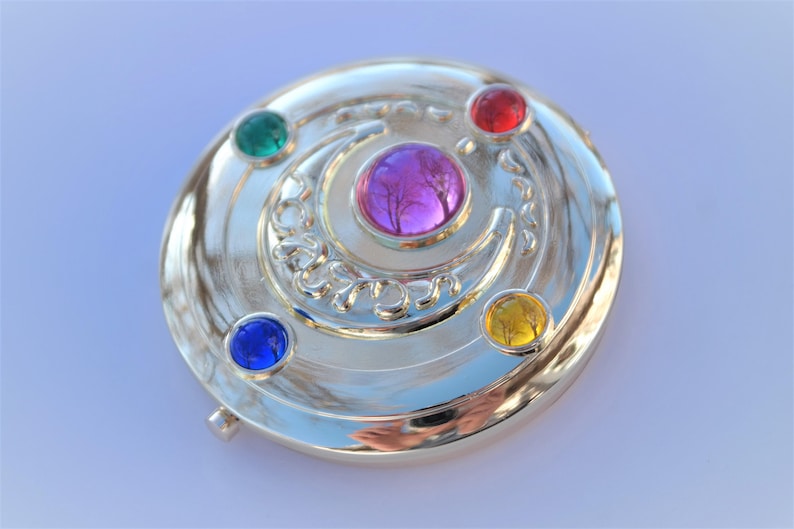 May include: A silver-colored compact mirror with a crescent moon design and five colorful gems. The gems are red, green, blue, yellow, and pink.