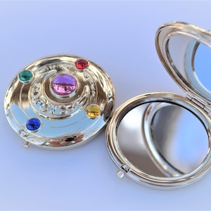 May include: A silver compact mirror with a decorative design featuring a crescent moon and five gemstones in red, yellow, green, blue, and purple. The mirror is open, revealing a reflective surface.