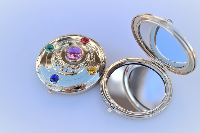 May include: A silver compact mirror with a decorative design featuring a crescent moon and five gemstones in red, yellow, green, blue, and purple. The mirror is open, revealing a reflective surface.