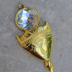 May include: A gold-colored pendant with a large, faceted, clear crystal at the top. The pendant has a shield-like shape with intricate etched designs and a small loop for hanging. The overall design is ornate and decorative.