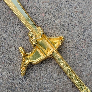 May include: A gold-colored sword with a decorative handle shaped like a dragon. The sword is lying on a gray surface.