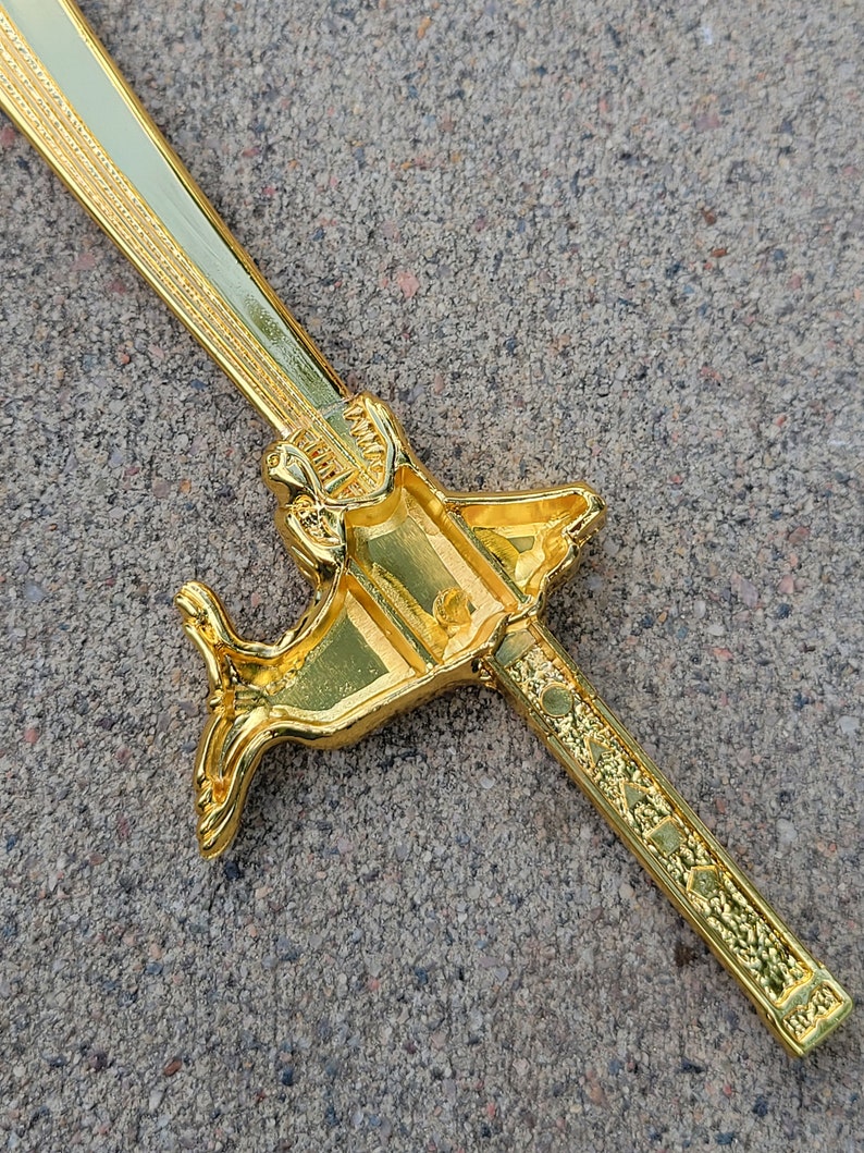 May include: A gold-colored sword with a decorative handle shaped like a dragon. The sword is lying on a gray surface.