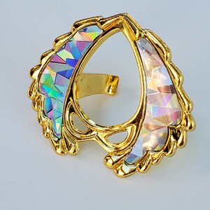 May include: A gold-colored ring with a pear-shaped opening and iridescent accents. The ring is decorated with a swirling, leaf-like design.