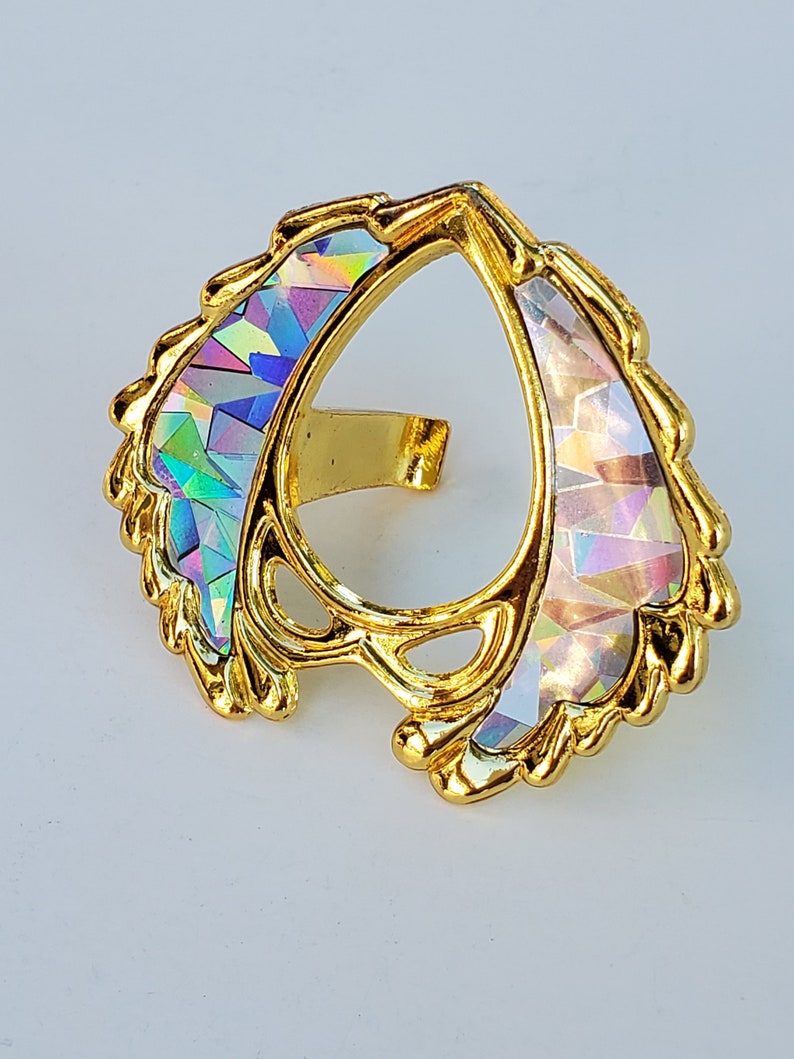 May include: A gold-colored ring with a pear-shaped opening and iridescent accents. The ring is decorated with a swirling, leaf-like design.