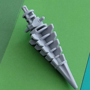 May include: A light gray plastic object with a pointed end and a series of angled, rectangular fins along its length. The top has a series of small, rectangular and cylindrical protrusions. The object is on a green surface.