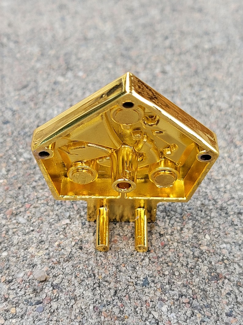 May include: Gold-colored metal object with a geometric shape and intricate details. The object has a hollow interior and appears to be a decorative piece.