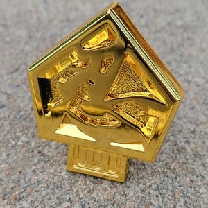 May include: A gold-colored metal pin shaped like a pentagon with a raised design inside. The pin has a small base with a series of lines.