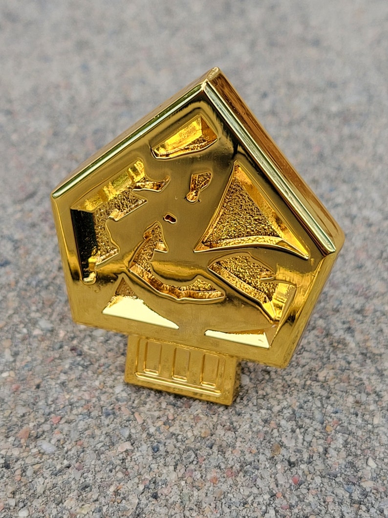 May include: A gold-colored metal pin shaped like a pentagon with a raised design inside. The pin has a small base with a series of lines.
