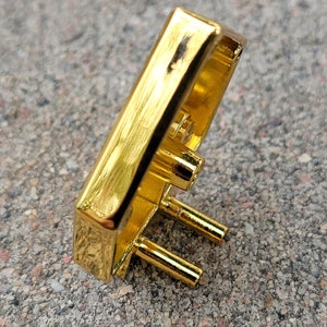 May include: Gold-colored metal buckle with a textured surface and three prongs. The buckle is rectangular in shape and has a slightly curved design.