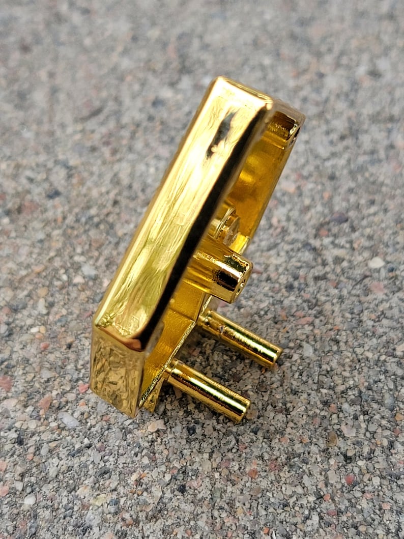 May include: Gold-colored metal buckle with a textured surface and three prongs. The buckle is rectangular in shape and has a slightly curved design.