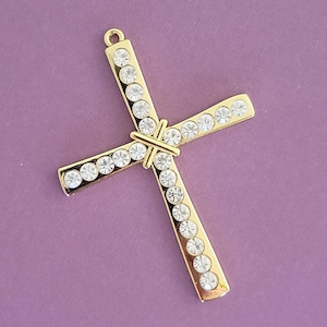 May include: Gold-colored cross pendant with clear, round rhinestones. The cross has a loop at the top for hanging and is set against a purple background. The rhinestones are evenly spaced along the arms of the cross.
