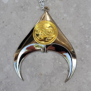 May include: A silver-toned pendant necklace with a gold-colored circular emblem at the center. The pendant has a crescent shape with a pointed top and a chain. The emblem features an ornate design.
